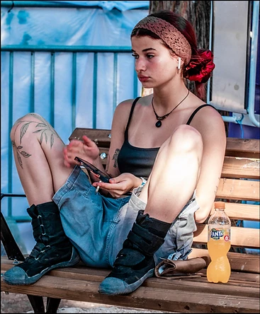 דיוקן דוקומנטרי צבעוני המציג צעירה יושבת על ספסל בעולם, כשהיא מחזיקה טלפון וסיגריה בידיה. הצילום שבוצע ב-Panasonic DMC-FZ1000 בתאורת יום טבעית, מתאפיין בחדות גבוהה על הדמות וביצירת קומפוזיציה המדגישה את המראה האורבני והקז'ואלי שלה, עם משחקי צבע של כחול, כתום ומרקמי עור וטקסטיל ברורים.
