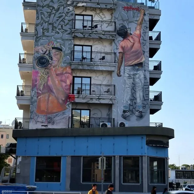 בתמונה נראה בניין מגורים רב-קומתי המעוטר בציור קיר (גרפיטי) אמנותי וגדול ממדים, המתאר דמויות אנושיות העוסקות בצילום. הקומפוזיציה מדגישה את הניגוד בין האמנות הסטטית שעל הקיר לבין התנועה הדינמית של הולכי הרגל והמכונית החולפת ברחוב. תאורת היום הטבעית מחמיאה לפרטים הצבעוניים שבציור ולמרקם הבטון של הבניין.