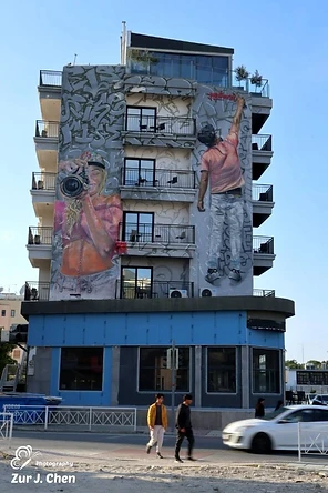 בתמונה נראה בניין מגורים רב-קומתי המעוטר בציור קיר (גרפיטי) אמנותי וגדול ממדים, המתאר דמויות אנושיות העוסקות בצילום. הקומפוזיציה מדגישה את הניגוד בין האמנות הסטטית שעל הקיר לבין התנועה הדינמית של הולכי הרגל והמכונית החולפת ברחוב. תאורת היום הטבעית מחמיאה לפרטים הצבעוניים שבציור ולמרקם הבטון של הבניין.