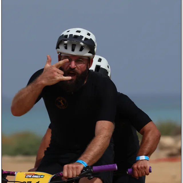 צילום ספורט דינמי המתעד שני רוכבים על אופני טנדם בחוף הים. הרוכב הקדמי בולט בהבעת פנים מלאת אנרגיה ותנועת יד, תוך שמירה על פוקוס חד וקומפוזיציה הדוקה. התאורה החזקה מבליטה את פרטי הקסדות והלבוש, בעוד עומק השדה הרדוד יוצר הפרדה יפה מהרקע המטושטש, ומעניק לתמונה תחושת תנועה ואווירה ספורטיבית אותנטית.