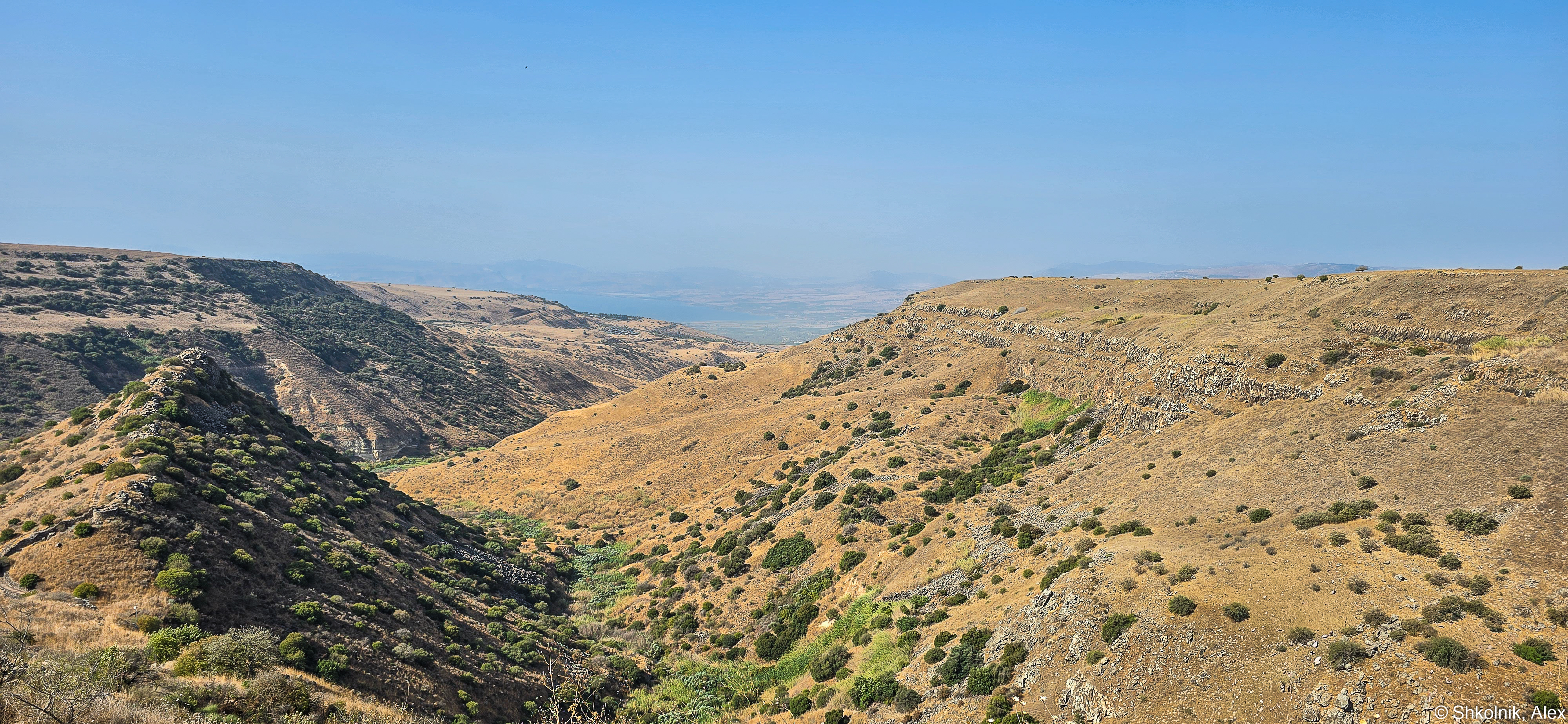 צילום נוף רחב ידיים המתאר ערוץ נחל עמוק המוקף בגבעות צחיחות למחצה, המעוטרות בצמחייה ים-תיכונית נמוכה. השמים הצלולים והתאורה העילית מדגישים את קווי המתאר הטופוגרפיים ואת המרקמים הסלעיים של המדרונות. הקומפוזיציה מנצלת את עומק השדה הרב כדי ליצור תחושת מרחב, כאשר הערוץ מוביל את העין אל עבר האופק הרחוק.