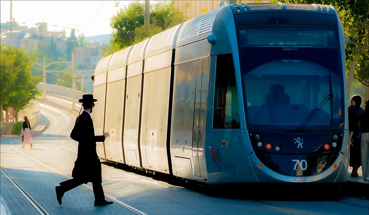 סצנה אורבנית דינמית המציגה גבר בלבוש חרדי חוצה מסילה בעולם, בעת שרכבת קלה מודרנית חולפת בסמוך. תאורת השמש האחורית יוצרת הילה רכה וקונטרסט גבוה, המדגיש את קווי המתאר של הרכבת והדמות. הקומפוזיציה משלבת תנועה אנושית אל מול המסה הסטטית והטכנולוגית של הקרון, תוך שמירה על עומק שדה רחב המאפשר הבנה של המרחב העירוני.