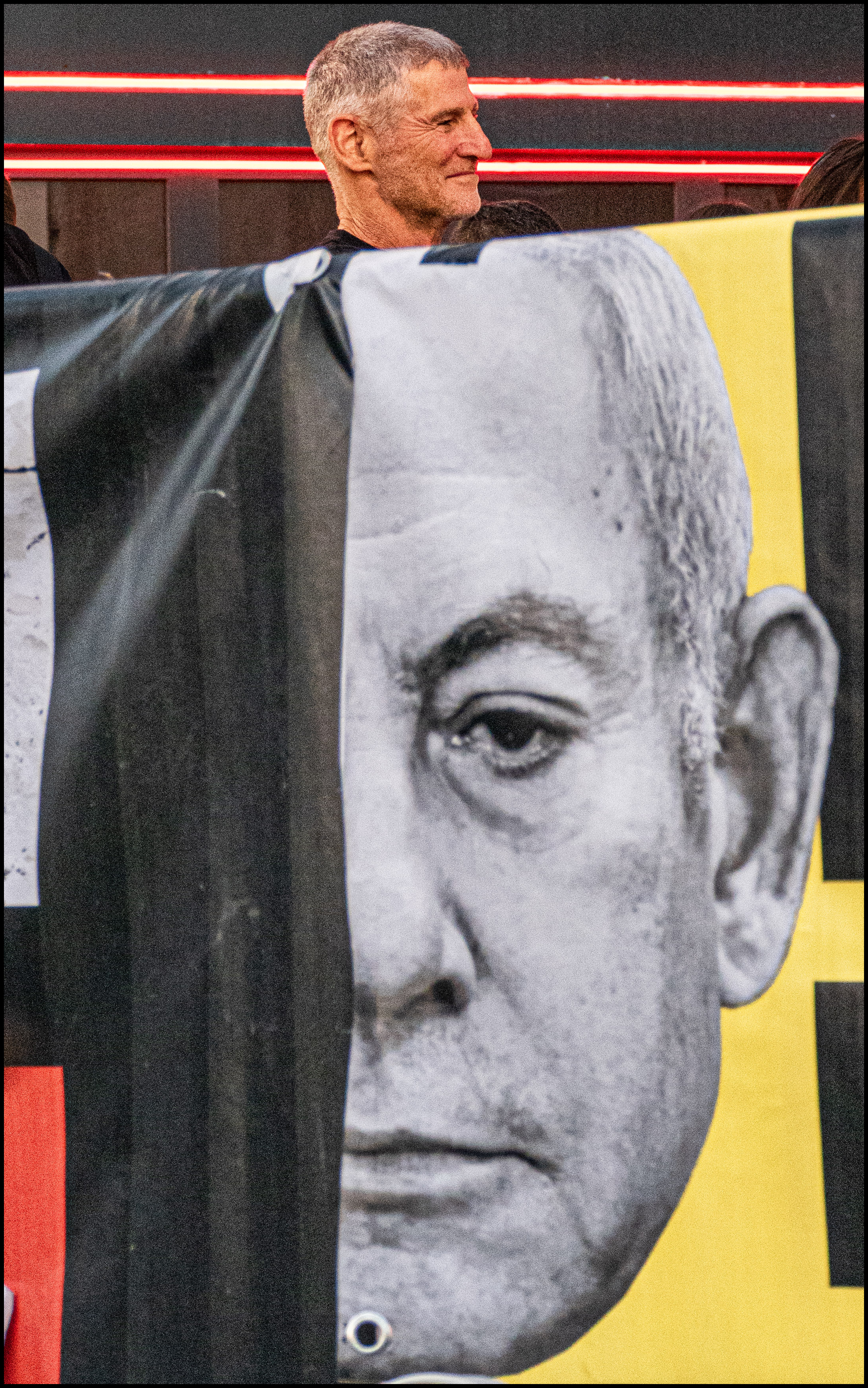 בתצלום דוקומנטרי עוצמתי שנלכד בעולם, נראה אדם העומד מאחורי כרזה פוליטית גדולה הנושאת דיוקן בשחור-לבן. השילוב בין פניו החיים של האדם לבין הדמות המודפסת יוצר משחק חזותי מעניין. הצילום משתמש בתאורה טבעית וקומפוזיציה חתוכה המדגישה את הבעת הפנים המרוכזת, תוך שמירה על ניגודיות גבוהה בצבעים ובמרקמים בין הבד לבין הדמות שמאחוריו.