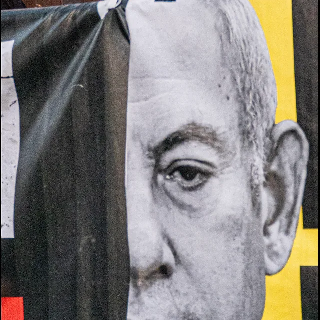 בתצלום דוקומנטרי עוצמתי שנלכד בעולם, נראה אדם העומד מאחורי כרזה פוליטית גדולה הנושאת דיוקן בשחור-לבן. השילוב בין פניו החיים של האדם לבין הדמות המודפסת יוצר משחק חזותי מעניין. הצילום משתמש בתאורה טבעית וקומפוזיציה חתוכה המדגישה את הבעת הפנים המרוכזת, תוך שמירה על ניגודיות גבוהה בצבעים ובמרקמים בין הבד לבין הדמות שמאחוריו.