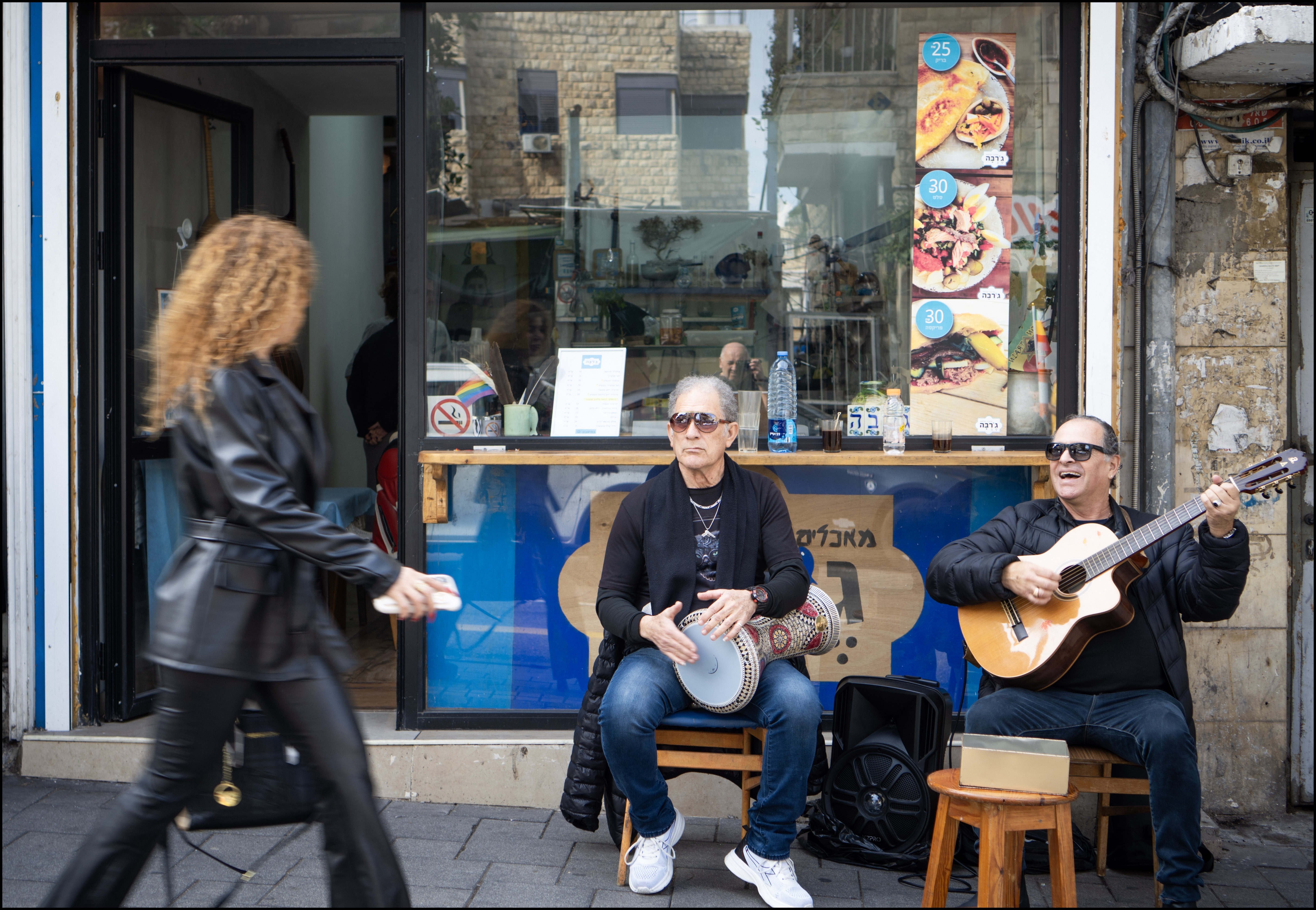 סצנת רחוב תוססת בעולם, המציגה שני מוזיקאים מנגנים מחוץ לחנות קטנה. בעוד האחד מנגן בגיטרה ושר והשני מלווה בדרבוקה, דמות מטושטשת חולפת על פניהם במהירות, מה שיוצר ניגודיות מעניינת בין הסטטיות של הנגנים לתנועה המהירה ברקע. הקומפוזיציה מנצלת את חזית החנות כרקע מעניין ומפורט, בעוד התאורה הטבעית מדגישה את הבעות הפנים של המוזיקאים.
