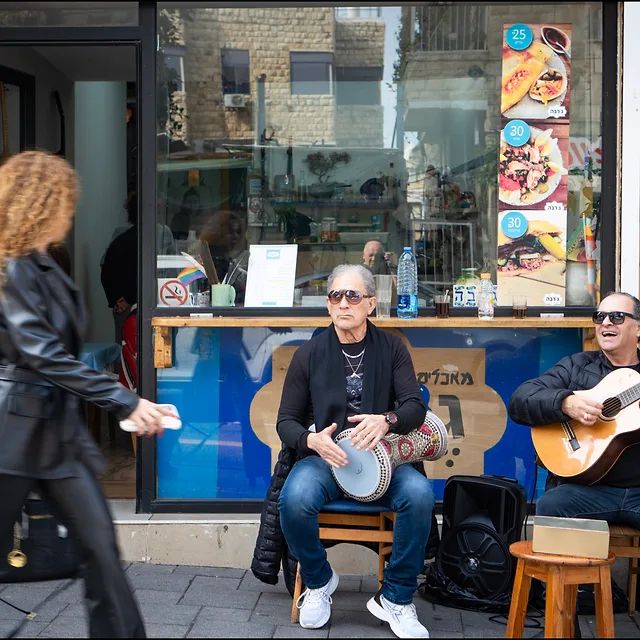 סצנת רחוב תוססת בעולם, המציגה שני מוזיקאים מנגנים מחוץ לחנות קטנה. בעוד האחד מנגן בגיטרה ושר והשני מלווה בדרבוקה, דמות מטושטשת חולפת על פניהם במהירות, מה שיוצר ניגודיות מעניינת בין הסטטיות של הנגנים לתנועה המהירה ברקע. הקומפוזיציה מנצלת את חזית החנות כרקע מעניין ומפורט, בעוד התאורה הטבעית מדגישה את הבעות הפנים של המוזיקאים.