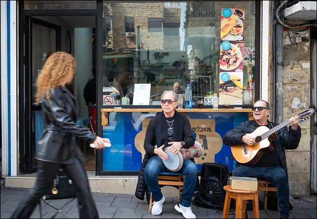 סצנת רחוב תוססת בעולם, המציגה שני מוזיקאים מנגנים מחוץ לחנות קטנה. בעוד האחד מנגן בגיטרה ושר והשני מלווה בדרבוקה, דמות מטושטשת חולפת על פניהם במהירות, מה שיוצר ניגודיות מעניינת בין הסטטיות של הנגנים לתנועה המהירה ברקע. הקומפוזיציה מנצלת את חזית החנות כרקע מעניין ומפורט, בעוד התאורה הטבעית מדגישה את הבעות הפנים של המוזיקאים.
