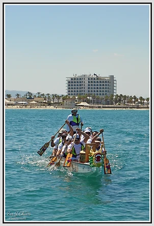 סירת דרקון עמוסה בחתירים נמרצים חוצה את המים הצלולים מול קו החוף של עכו, כאשר דמות ראש דרקון דמיוני מעטרת את חרטומה. התאורה הבהירה מדגישה את תנועת המשוטים והשובל שיוצרת הסירה, בעוד שהקומפוזיציה המרכזית ממקמת את הקבוצה אל מול מבנה המלון המרוחק, ומעבירה תחושת פעילות ספורטיבית אנרגטית ביום שטוף שמש.