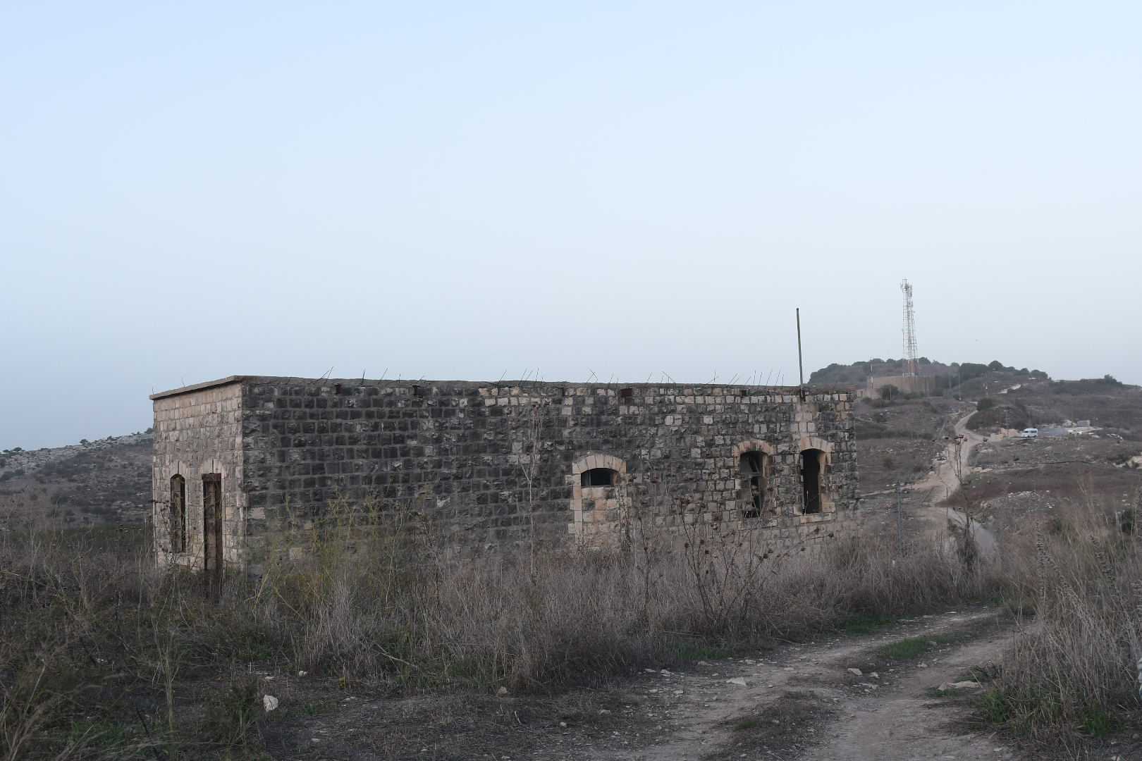 צילום נוף המציג מבנה אבן עתיק ונטוש הניצב בלב שטח פתוח וצחיח למחצה. קומפוזיציית הצילום משלבת בין המבנה לדרך עפר מפותלת המובילה אל עבר אופק רחוק, תחת שמיים בהירים ושטוחים. שימוש בעדשה רחבה (18 מ"מ) מדגיש את תחושת המרחב, בעוד תאורת היום הרכה מדגישה את הטקסטורה המחוספסת של האבנים הישנות.