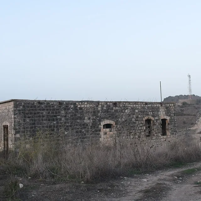 צילום נוף המציג מבנה אבן עתיק ונטוש הניצב בלב שטח פתוח וצחיח למחצה. קומפוזיציית הצילום משלבת בין המבנה לדרך עפר מפותלת המובילה אל עבר אופק רחוק, תחת שמיים בהירים ושטוחים. שימוש בעדשה רחבה (18 מ"מ) מדגיש את תחושת המרחב, בעוד תאורת היום הרכה מדגישה את הטקסטורה המחוספסת של האבנים הישנות.