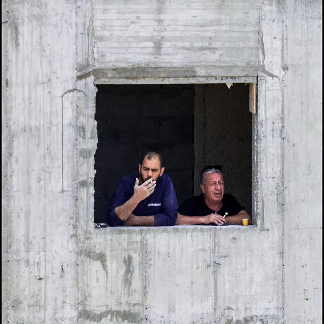 שני גברים מבוגרים נשקפים מחלון של מבנה בבנייה גולמית בעולם, כשהם נחים לרגע במהלך יום עבודה. הצילום הממוקד משתמש בטקסטורות המחוספסות של הבטון החשוף כמסגרת טבעית המדגישה את הדמויות. חשיפה קצרה של 1/1000 שנ' תרמה לחדות גבוהה, בעוד עומק השדה הממוצע שומר על הפרטים בחלון במיקוד ברור אל מול הרקע.