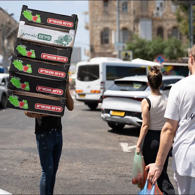 צילום רחוב דינמי בעולם, המתעד רגע יומיומי שבו אדם נושא ערימת ארגזי פירות על ראשו בלב המולה עירונית. השימוש בעדשת 35 מ"מ ומפתח צמצם F5.6 יצר עומק שדה המשלב בין הדמות המרכזית לבין הסביבה, תוך שמירה על תאורת יום חדה המדגישה את הניגודיות שבין עבודת הכפיים לבין עוברי האורח מסביב.