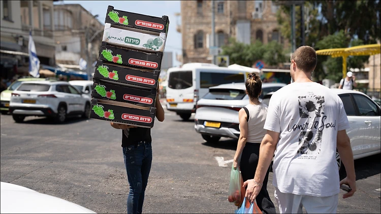 צילום רחוב דינמי בעולם, המתעד רגע יומיומי שבו אדם נושא ערימת ארגזי פירות על ראשו בלב המולה עירונית. השימוש בעדשת 35 מ"מ ומפתח צמצם F5.6 יצר עומק שדה המשלב בין הדמות המרכזית לבין הסביבה, תוך שמירה על תאורת יום חדה המדגישה את הניגודיות שבין עבודת הכפיים לבין עוברי האורח מסביב.