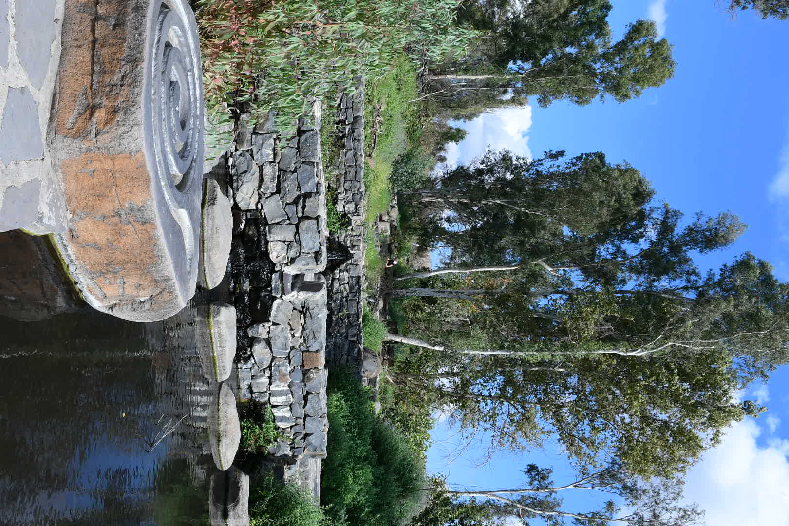 צילום נוף שליו ופסטורלי המציג פלג מים רגוע עם אבני מדרך חוצות, הנשפך למבנה אבן עתיק למראה. השימוש בעדשה רחבה (18 מ"מ) וצמצם סגור (f/11) מבטיחים עומק שדה רב וחדות מקסימלית מהקרוב לרחוק. השמיים הכחולים והצמחייה העבותה מעניקים תחושת טבע רעננה, בעוד קווי המתאר המעוגלים של האבן הקדמית מוסיפים עניין קומפוזיציוני.