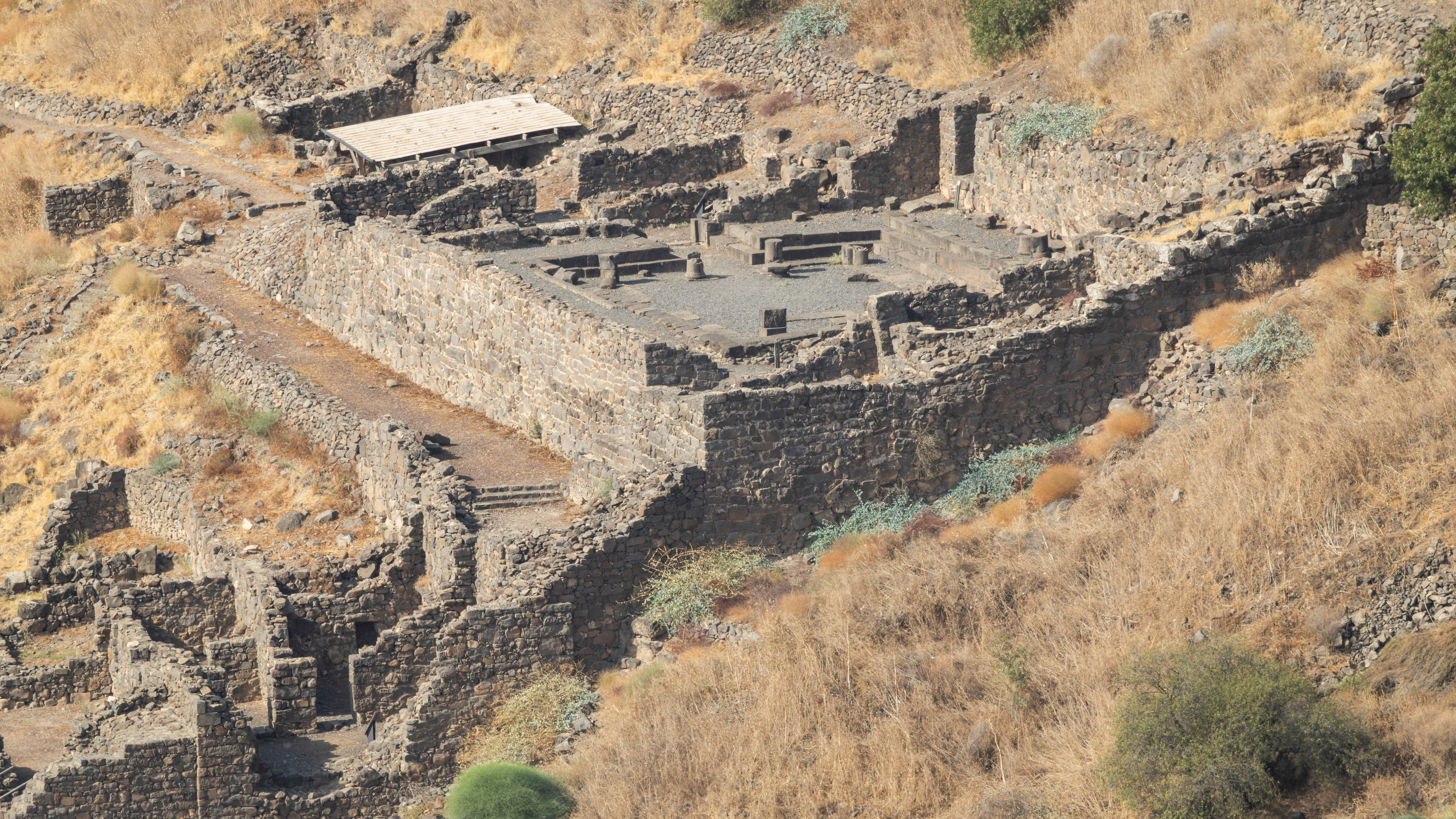 צילום אווירי מרשים של אתר עתיקות בנוי מאבני בזלת כהות, המשתלב במדרון הררי צהבהב ויבש. השימוש בעדשת טלפוטו (661 מ"מ) בצירוף מפתח צמצם F/10, יצר עומק שדה עמוק ששמר על חדות גבוהה בכל חלקי המבנה הארכיאולוגי, תוך הדגשת הטקסטורות המחוספסות של האבנים והצמחייה האופיינית לאזור.