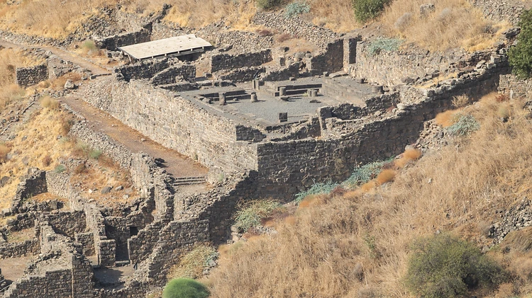 צילום אווירי מרשים של אתר עתיקות בנוי מאבני בזלת כהות, המשתלב במדרון הררי צהבהב ויבש. השימוש בעדשת טלפוטו (661 מ"מ) בצירוף מפתח צמצם F/10, יצר עומק שדה עמוק ששמר על חדות גבוהה בכל חלקי המבנה הארכיאולוגי, תוך הדגשת הטקסטורות המחוספסות של האבנים והצמחייה האופיינית לאזור.