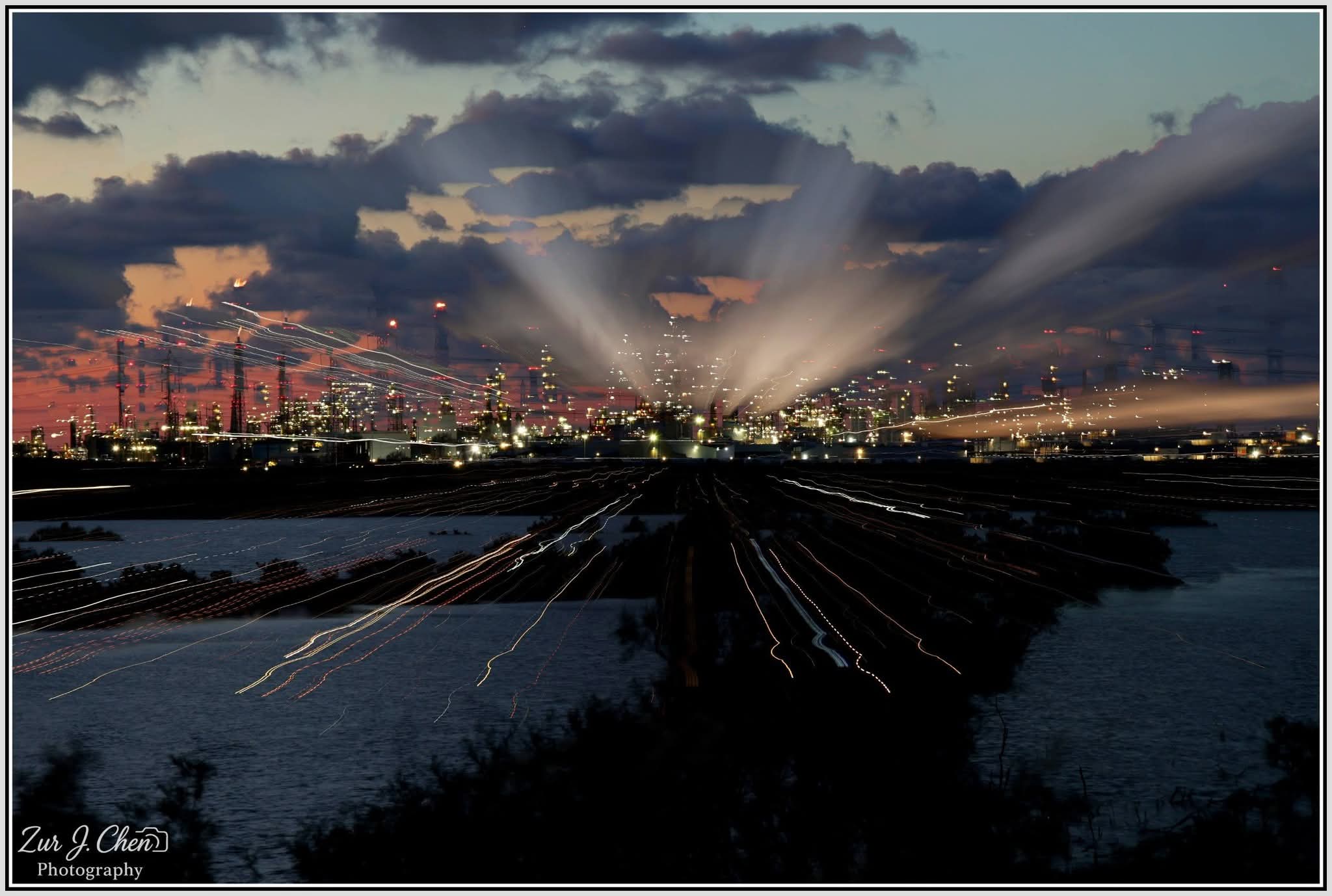 צילום חשיפה ארוכה מרתק של מתחם תעשייתי לילי, המציג משחקי אורות ופסי תנועה היוצרים אפקט דינמי מעל גוף מים. השילוב בין אורות המפעל, עשן המשתקף באור וצבעי הדמדומים העמוקים בשמיים, יוצר אווירה תעשייתית דרמטית וייחודית. הקומפוזיציה רחבה, עם דגש על הקווים המוליכים שנוצרו מהתנועה ועל הניגודיות שבין הטבע האפל לחום האור המלאכותי.