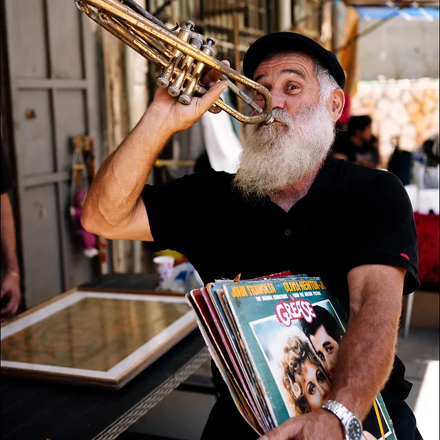 צילום דוקומנטרי המציג אדם בעל זקן לבן מרשים, מנגן בחצוצרה בעודו אוחז בערימת תקליטים וינטג' ברחובות העולם. השימוש בעדשת 35 מ"מ ומפתח צמצם של 2.8 יצר עומק שדה רך המבליט את הדמות על פני הרקע, תוך לכידת רגע אותנטי וצבעוני בתאורת יום טבעית וחדות גבוהה על פניו של האמן.