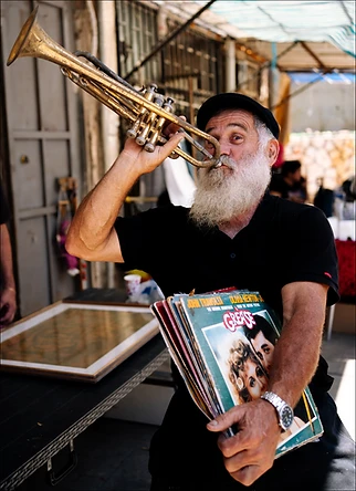 צילום דוקומנטרי המציג אדם בעל זקן לבן מרשים, מנגן בחצוצרה בעודו אוחז בערימת תקליטים וינטג' ברחובות העולם. השימוש בעדשת 35 מ"מ ומפתח צמצם של 2.8 יצר עומק שדה רך המבליט את הדמות על פני הרקע, תוך לכידת רגע אותנטי וצבעוני בתאורת יום טבעית וחדות גבוהה על פניו של האמן.