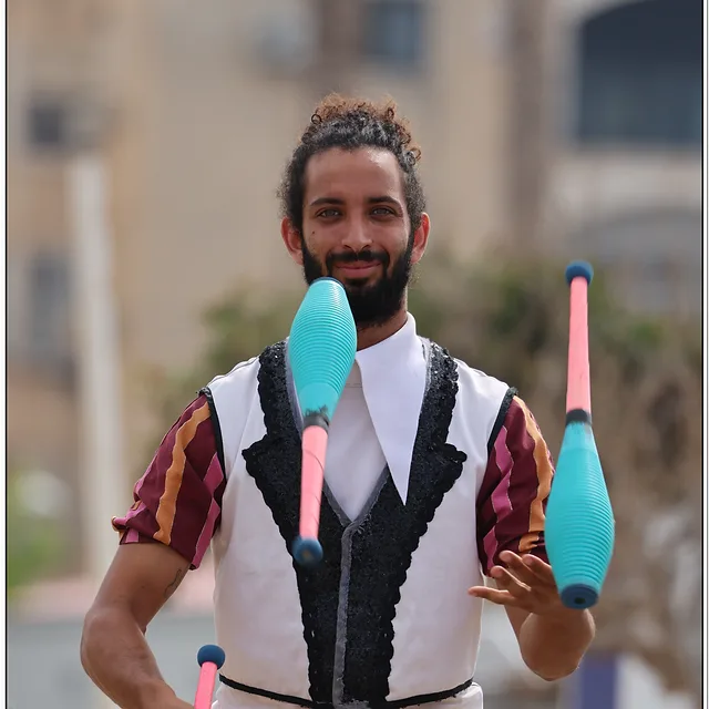 צילום דוקומנטרי של להטוטן המבצע מופע רחוב דינמי. האובייקט המרכזי ממוקם במרכז הפריים, כשהוא אוחז ומלהטט באלות צבעוניות בגווני טורקיז וורוד. טשטוש הרקע בזכות עומק שדה רדוד מדגיש את הפורטרט ואת הבעת פניו הממוקדת של האמן, בעוד התאורה הרכה מחמיאה לפרטי התלבושת ומבליטה את תנועת האלות באוויר.