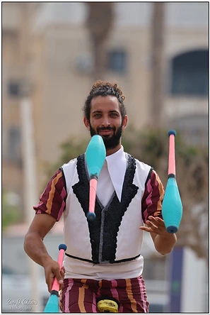 צילום דוקומנטרי של להטוטן המבצע מופע רחוב דינמי. האובייקט המרכזי ממוקם במרכז הפריים, כשהוא אוחז ומלהטט באלות צבעוניות בגווני טורקיז וורוד. טשטוש הרקע בזכות עומק שדה רדוד מדגיש את הפורטרט ואת הבעת פניו הממוקדת של האמן, בעוד התאורה הרכה מחמיאה לפרטי התלבושת ומבליטה את תנועת האלות באוויר.