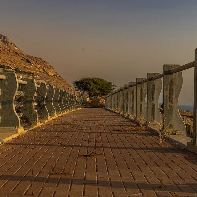 בתמונה נראה שביל מרוצף רחב המובל אל עבר האופק, מתוחם על ידי גדר בטון מודרנית עם עיצוב גלי. זווית הצילום הנמוכה יוצרת הולכת עין חזקה לעומק הפריים. אור השמש בשעה זו מעניק גוונים חמים לקרקע, בעוד המבנה ההררי והעץ המרוחק משלימים תחושת שלווה במרחב פתוח וחשוף.