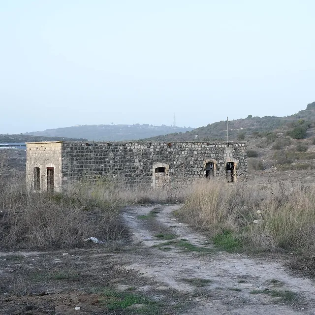 צילום נוף המציג מבנה אבן עתיק ונטוש הניצב בלב שטח פתוח וצחיח. הקומפוזיציה הרחבה, שצולמה במוקד 26 מ"מ, מדגישה את הבדידות של המבנה על רקע גבעות מרוחקות. התאורה הטבעית הרכה מעניקה למבנה מראה היסטורי, בעוד הצבעוניות המונוכרומטית והטקסטורה של אבני הבנייה יוצרות אווירה דוקומנטרית שקטה ונוסטלגית.