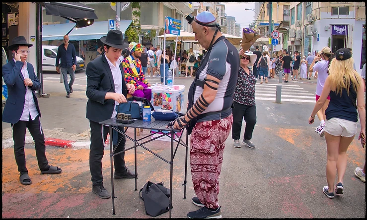 תצלום רחוב תוסס המציג סיטואציה יומיומית בעולם, שבו דמויות לבושות בבגדים שונים מתקבצות סביב שולחן קטן. השימוש בצמצם f/4.5 יצר עומק שדה המפריד מעט את הדמויות מהרקע העמוס, בעוד התאורה הטבעית והרכה מאפשרת הבחנה בפרטים ובמרקמים השונים של לבוש האנשים, החל בחליפות ועד למכנסיים צבעוניים ותפילין, המעניקים לצילום אופי דוקומנטרי עשיר.