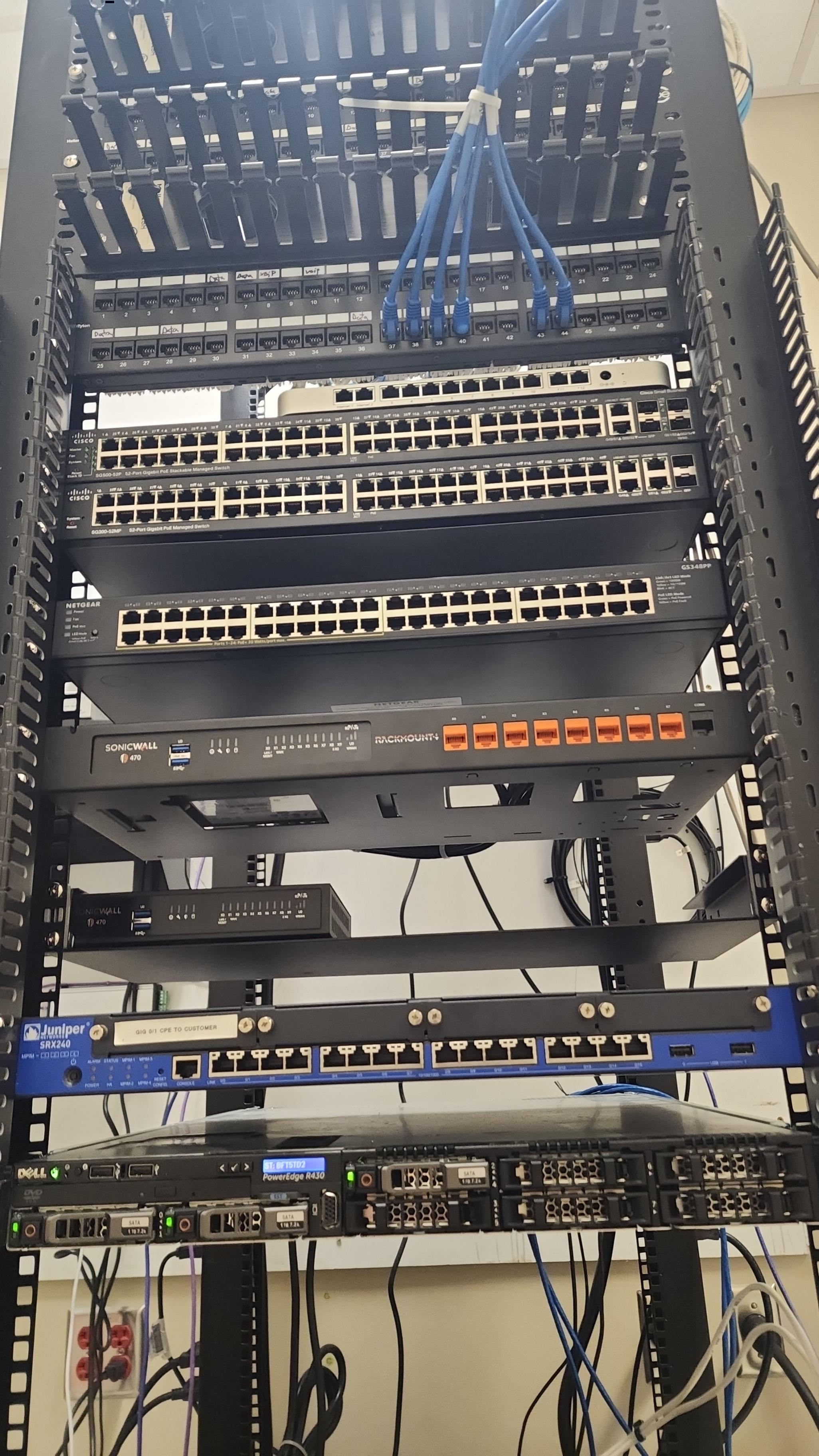 Network rack after cleanup with neatly dressed white patch cables, cable management channels, Cisco and Netgear switches, SonicWall firewall, Juniper router, and Dell server