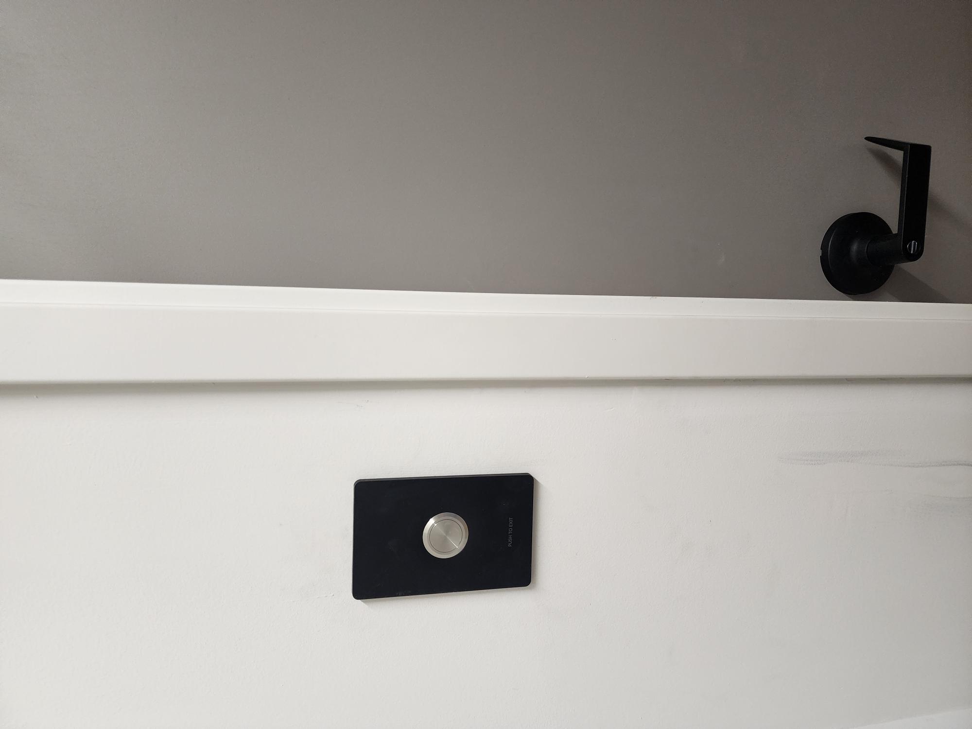 Black rectangular push-to-exit button with a brushed metal circle mounted on a white wall above a grey door frame