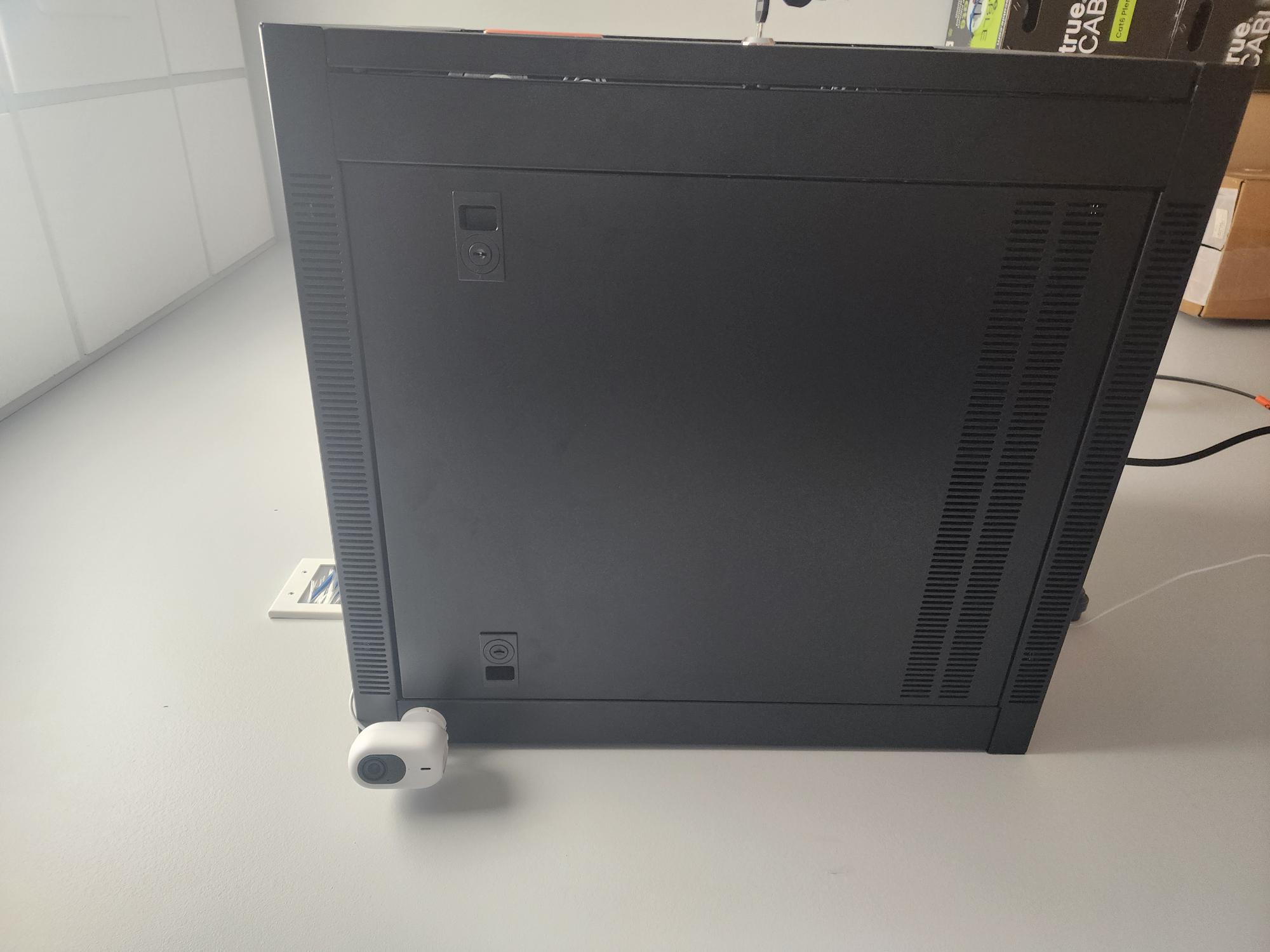 Small white security camera mounted to the side of a black server rack on a light grey floor