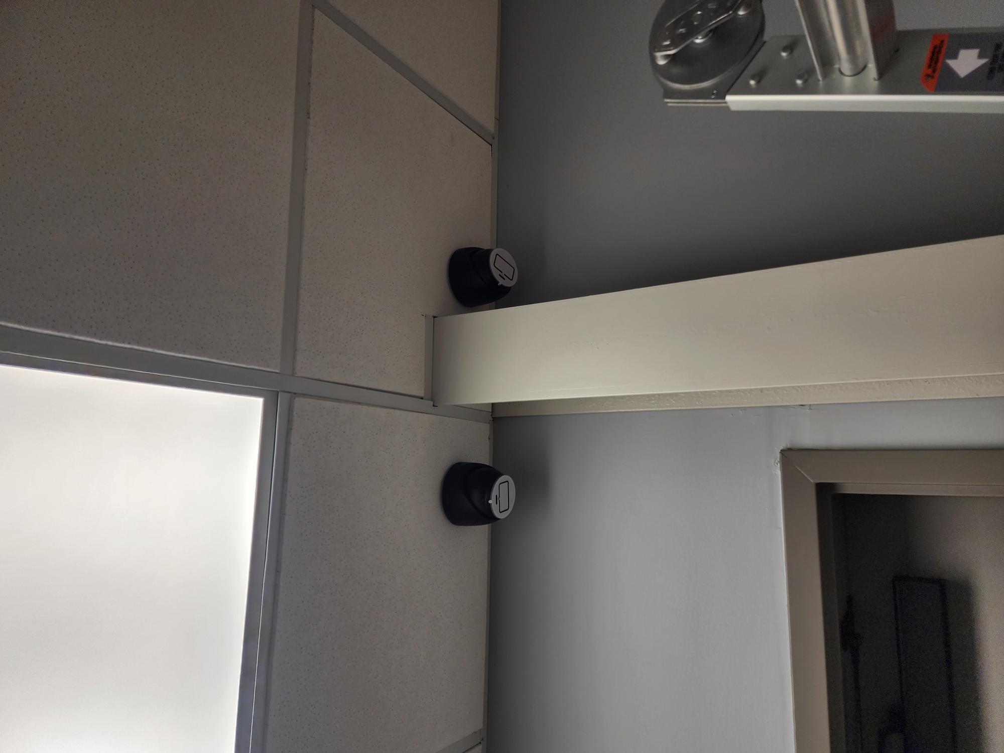 Two black low-voltage mounting plates with white faceplate inserts mounted vertically on a grey wall at a ceiling junction