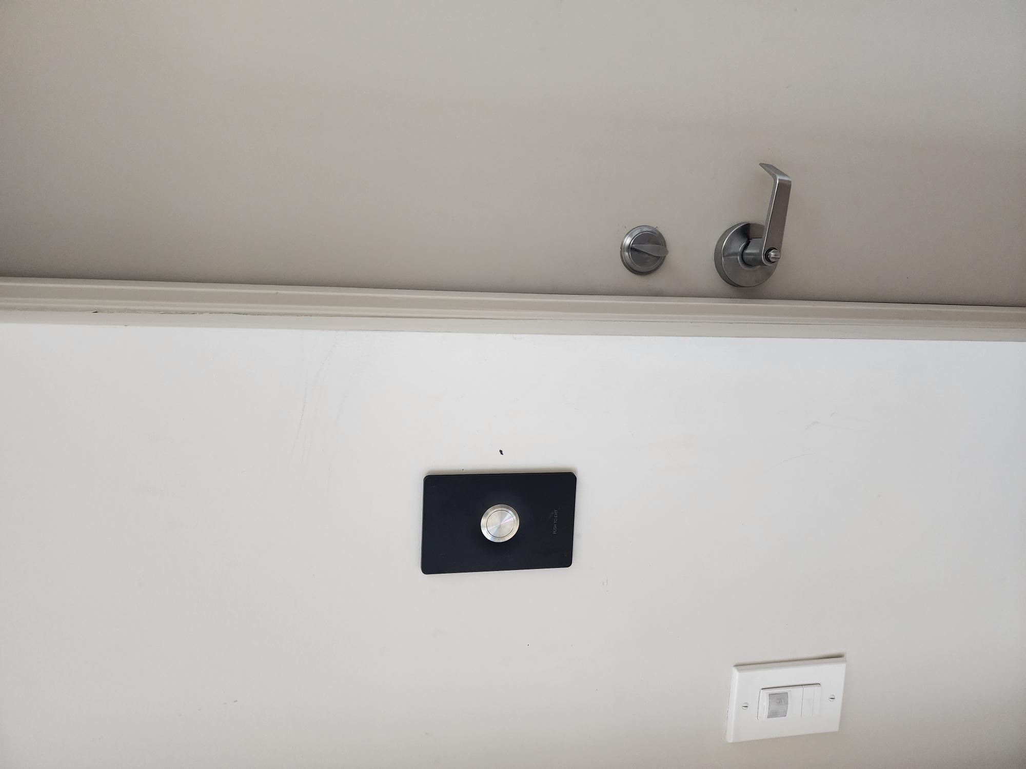 Push-to-exit button on a white wall with a door lever and a light switch mounted nearby