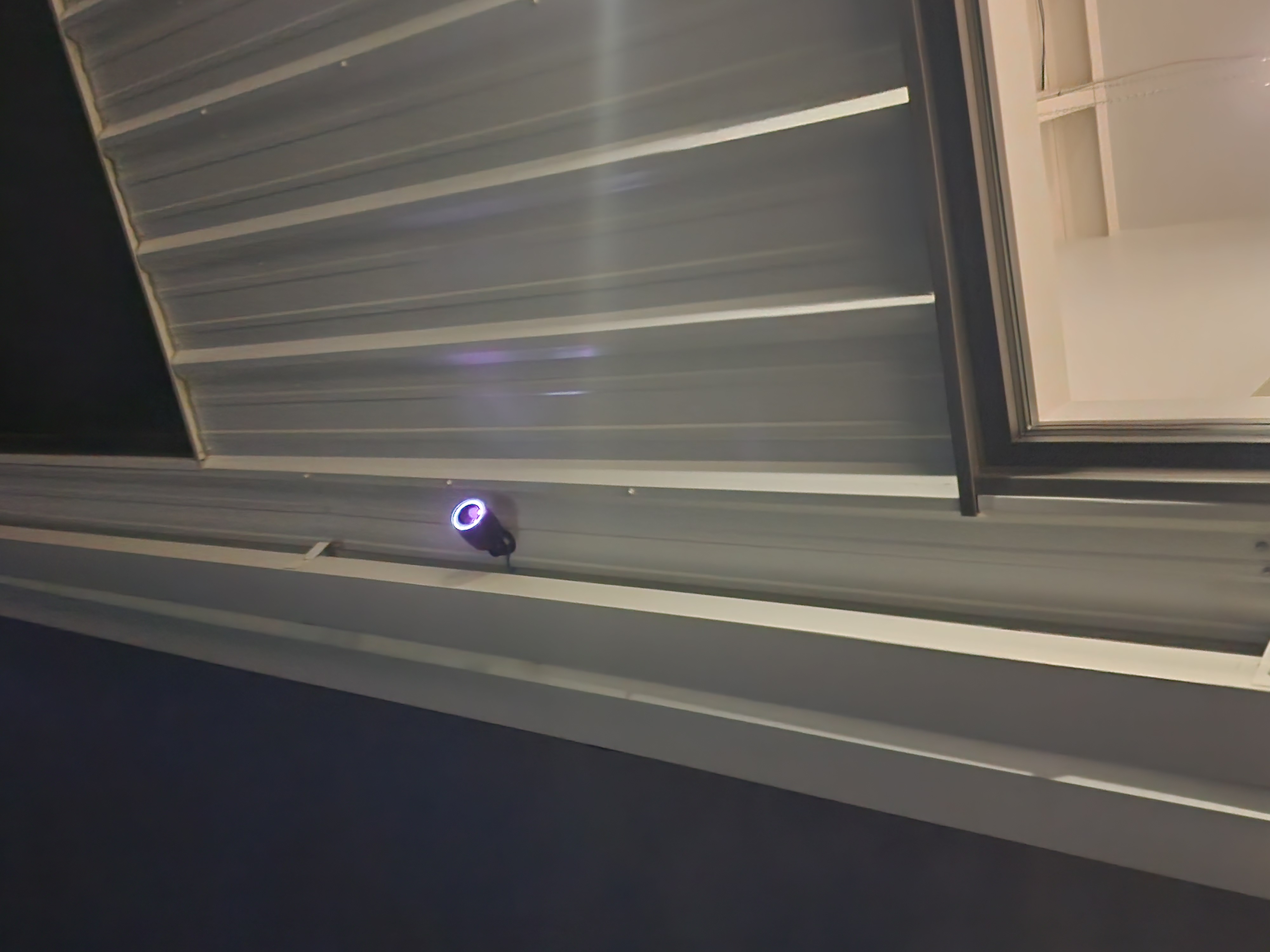 Black cylindrical security camera mounted under a white corrugated metal roof overhang with a glowing blue-violet ring around the lens