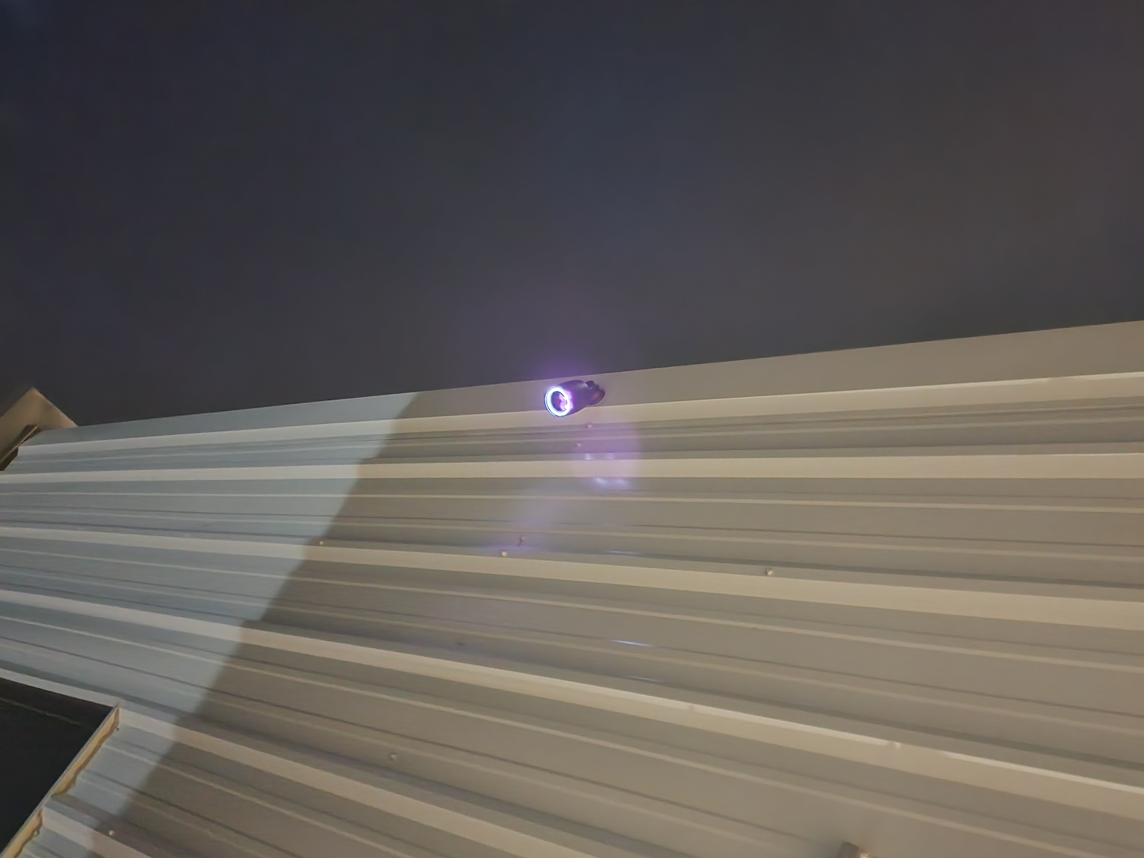 Black cylindrical security camera mounted on corrugated metal siding at night with a glowing LED ring around the lens