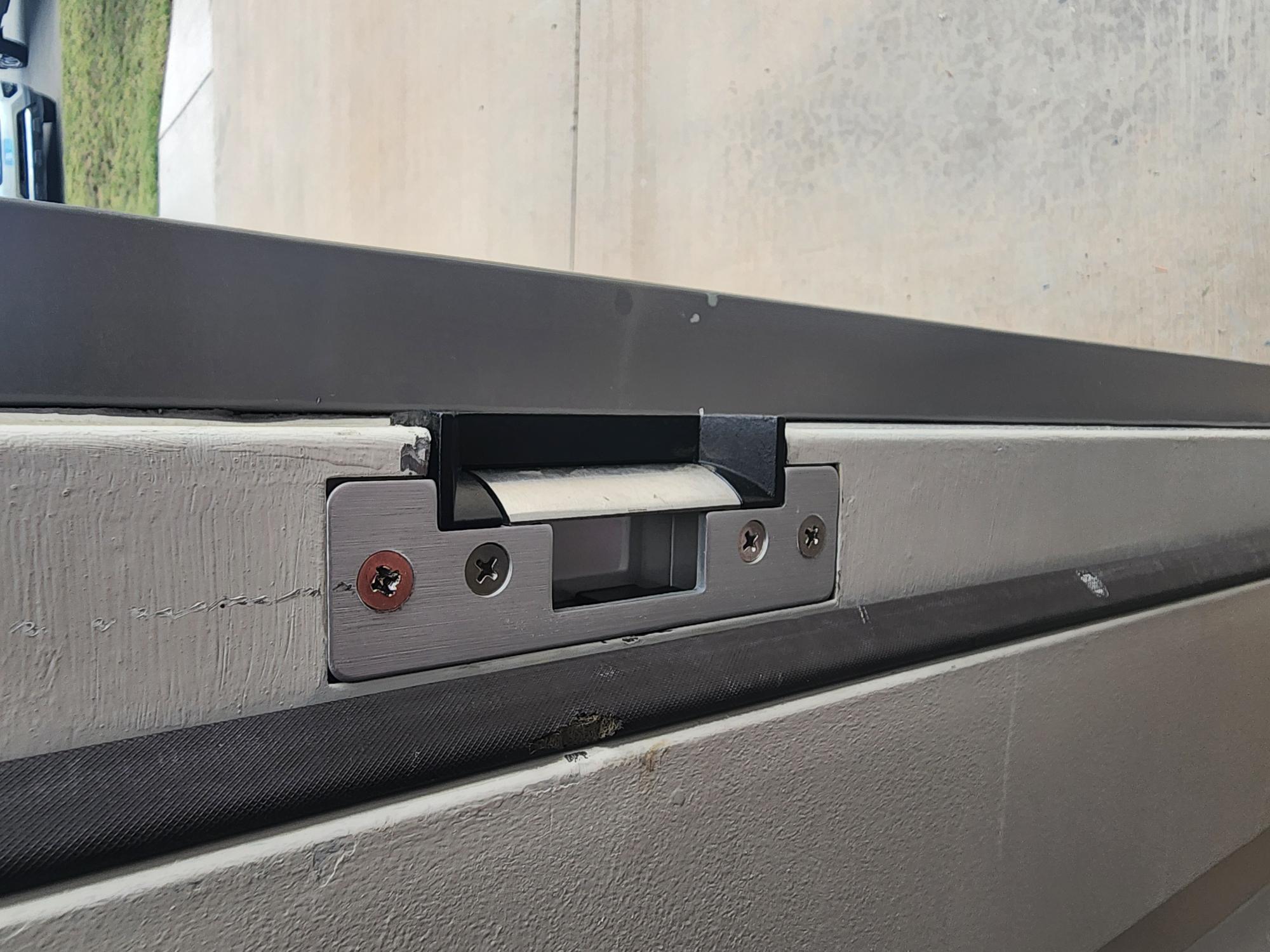 Close-up of an electric door strike flush-mounted into a grey door frame with weather stripping along the edge