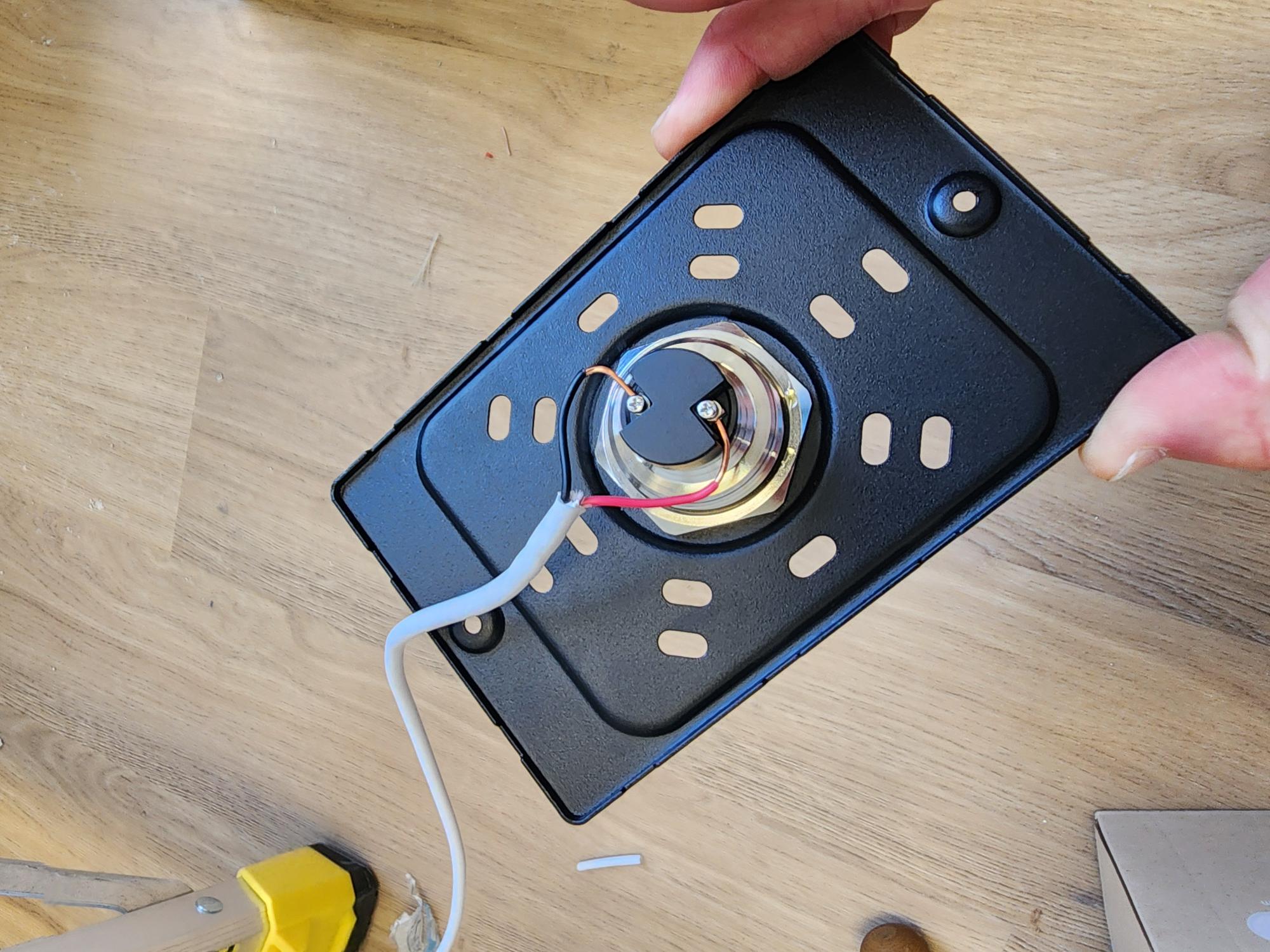 Hands holding a black metal bracket with a silver cylindrical lock actuator wired with red and black wires through a white cable