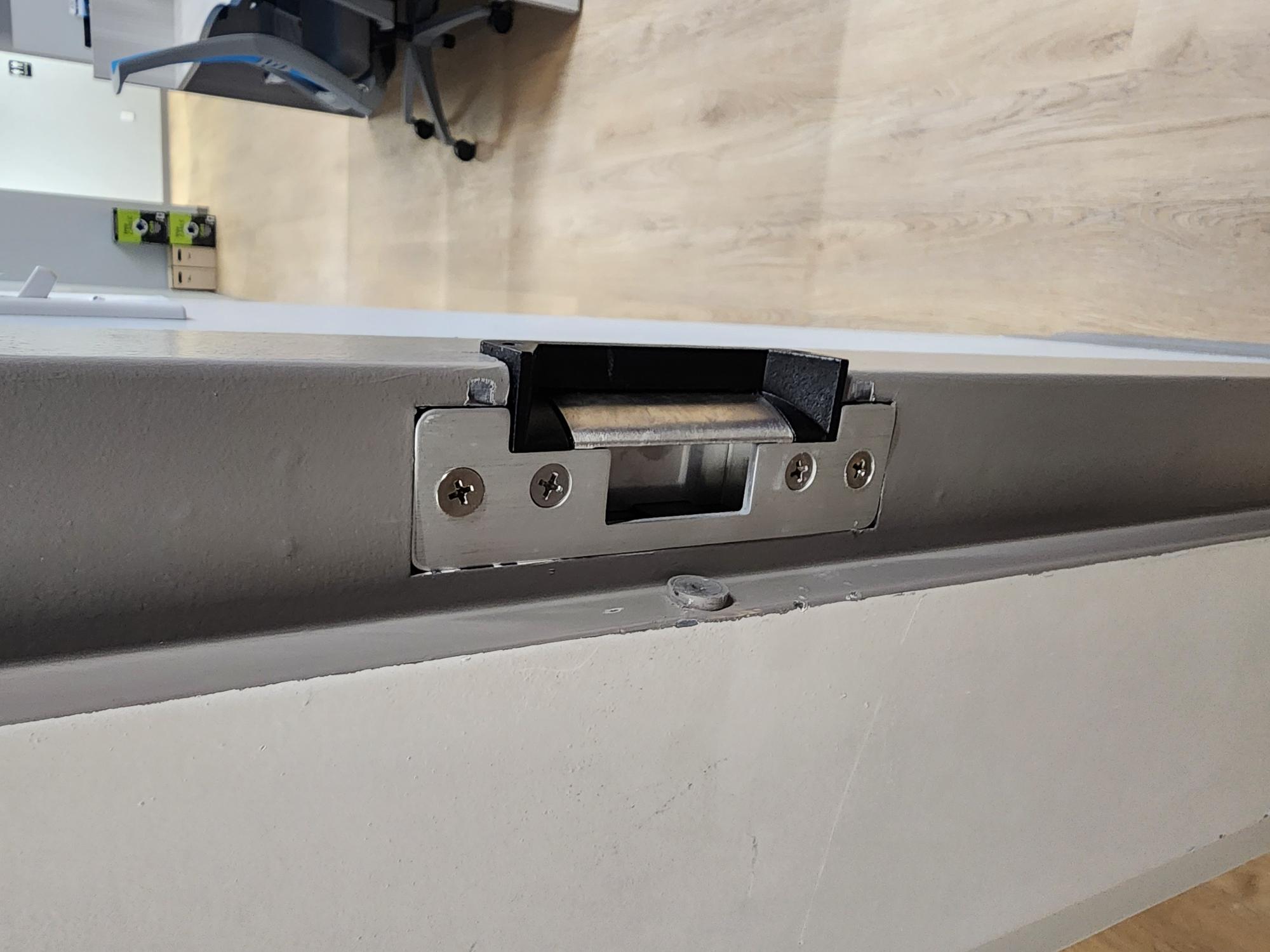 Electric strike plate mortised into a grey door frame with a push-to-exit button mounted on the wall nearby