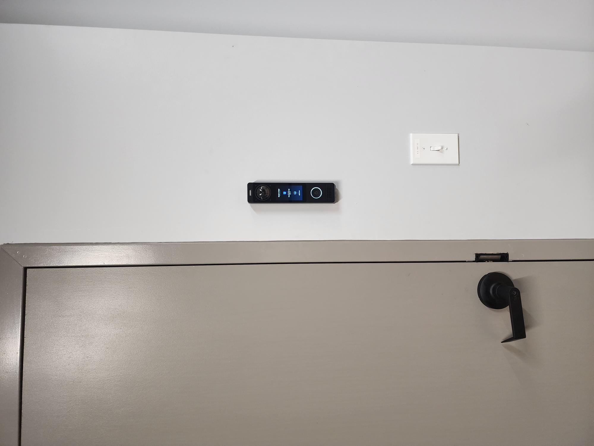 UniFi access control reader mounted on a wall above a grey door frame, showing a ready-to-adopt display
