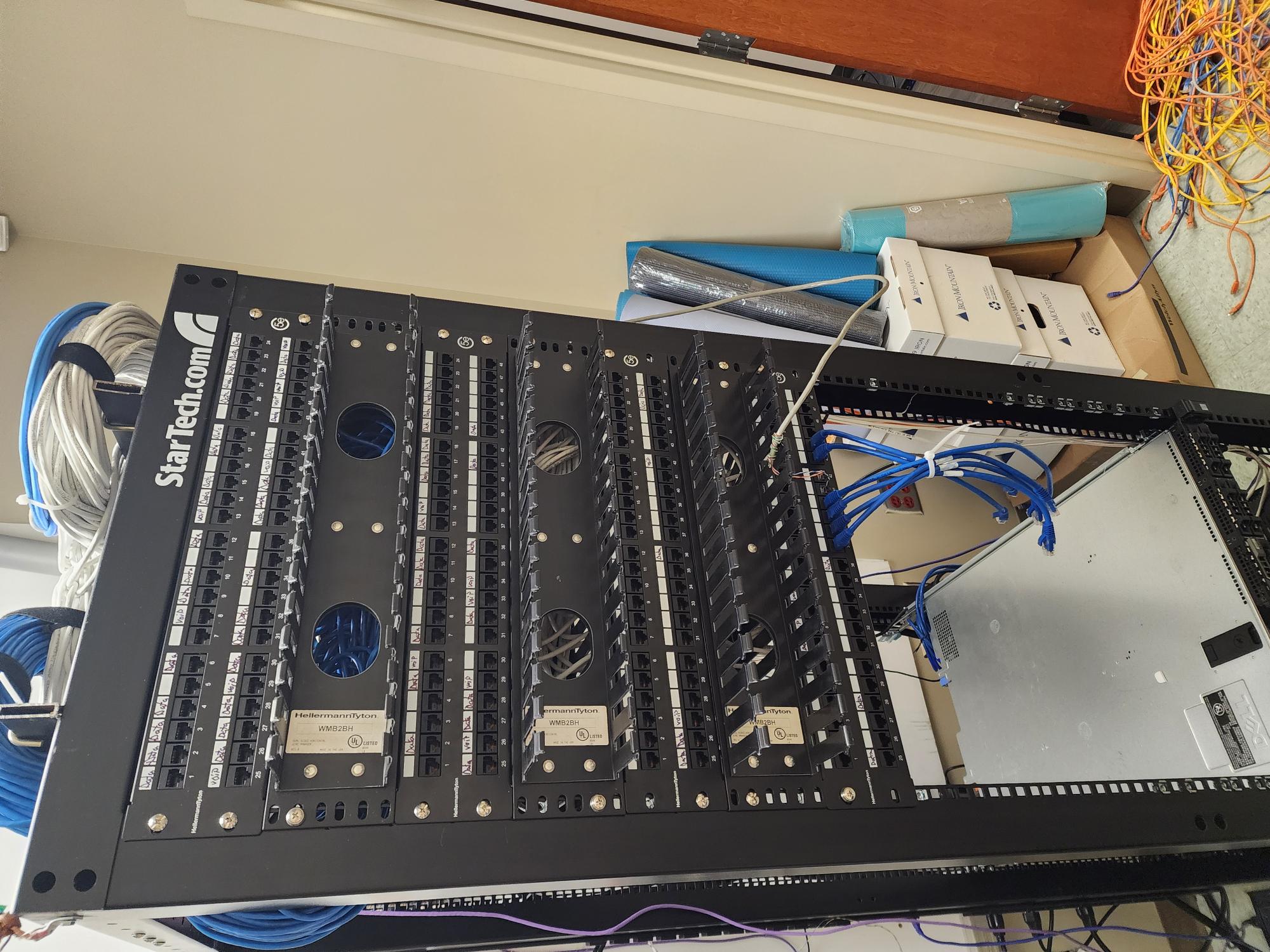 Rack of StarTech and HarmonyPlus patch panels with blue network cables before cleanup, with a server partially visible to the side