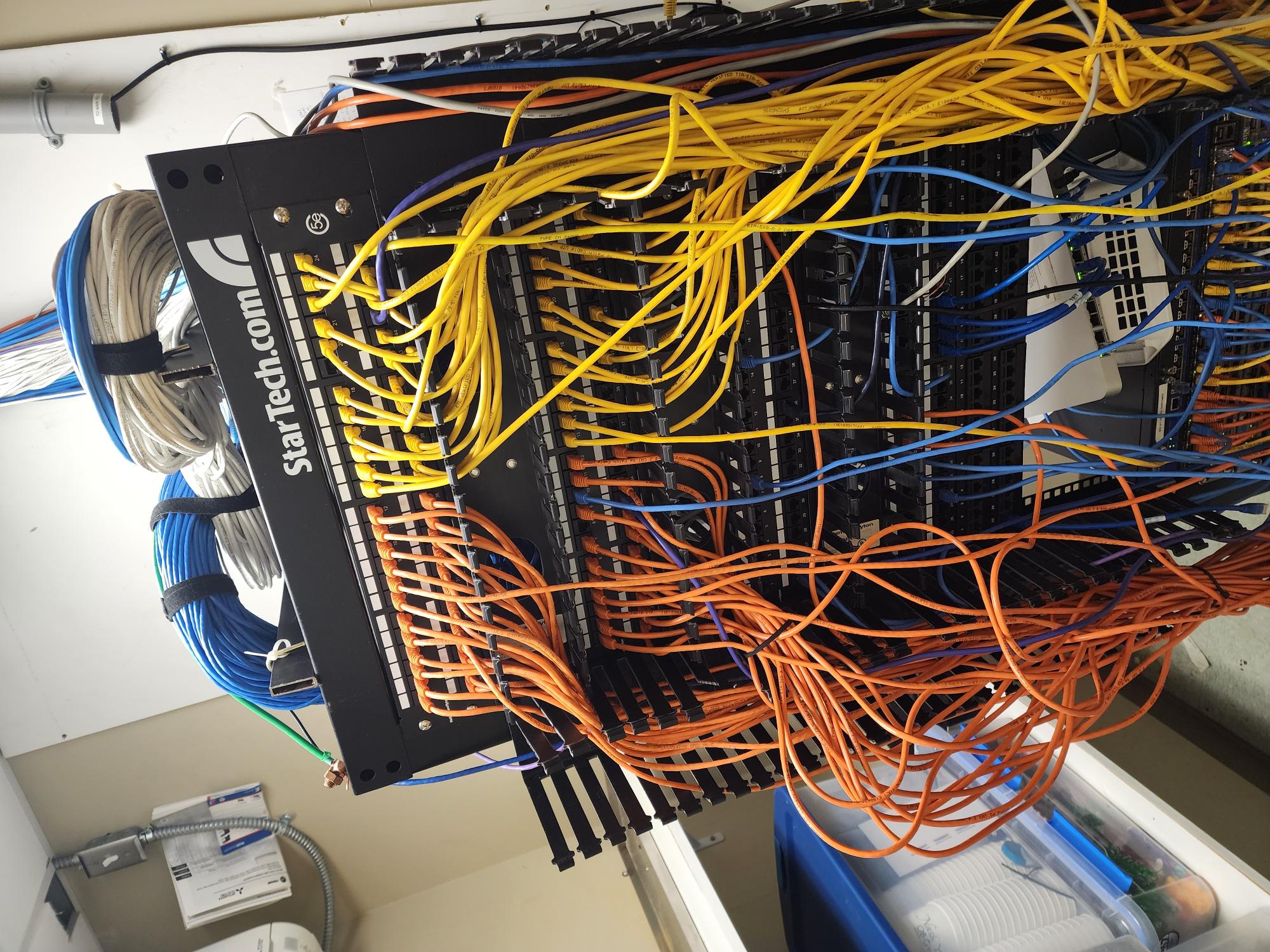 StarTech patch panel before cleanup, crowded with tangled yellow, blue, and orange patch cables draping over a cable management channel