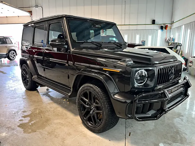 Black Mercedes-Benz G-Class viewed from the front quarter inside a garage, with its grille, round headlights and black wheels visible.