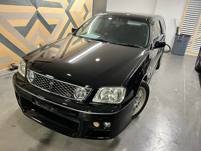 Black station wagon viewed from the front in a workshop beside a geometric wood-panel wall.