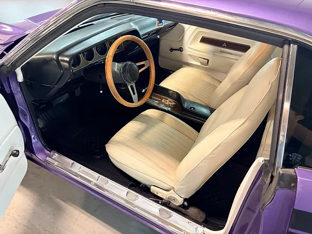 Cream seats and a wooden steering wheel inside a purple classic car, viewed through the open driver’s door.