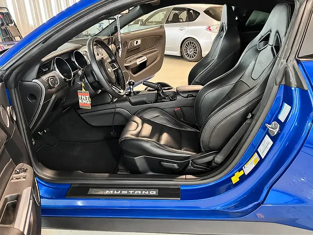 Blue Ford Mustang interior viewed through the open driver’s door, showing black leather sport seats, steering wheel, manual gear shifter, and carpets.