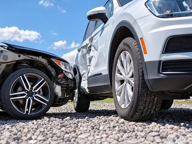 Two collision-damaged vehicles on gravel under a blue sky, showing a black car with front-end damage beside a white SUV with dented side doors.