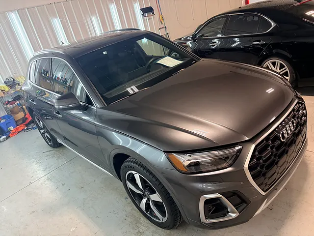 Grey Audi SUV viewed from the front quarter inside a workshop, with a black sedan behind it.