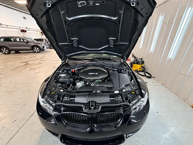 Black BMW M3 in a workshop with its hood raised, showing the V8 engine bay and front grille.