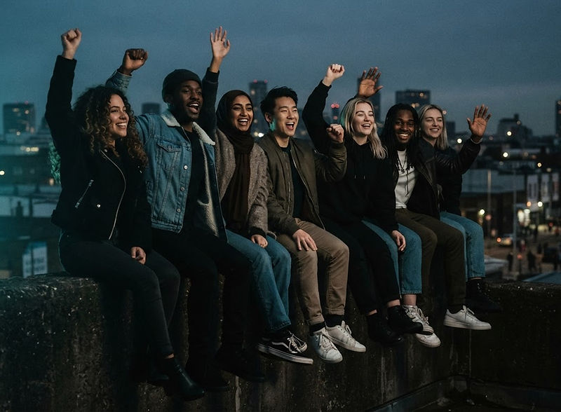 A diverse group of people sitting together on a dark wall, hands raised in solidarity