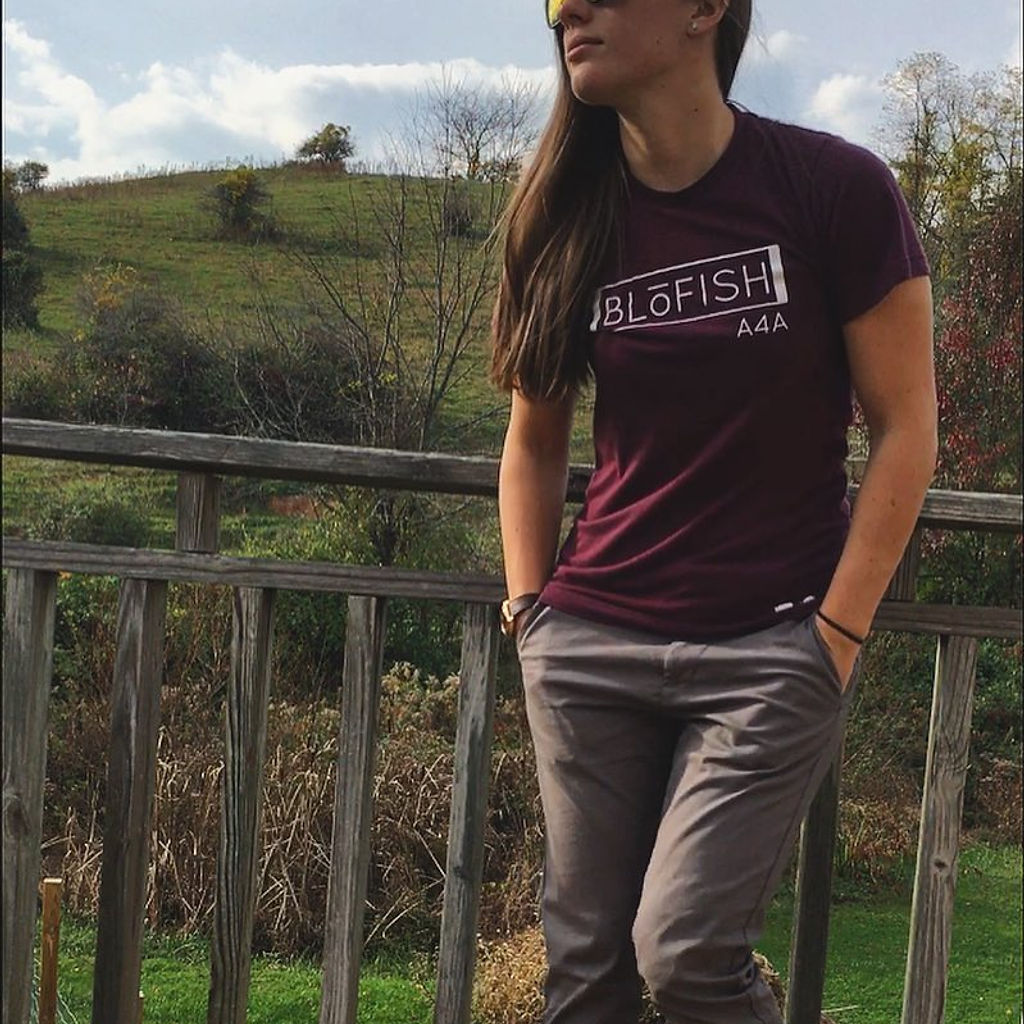 Lacie leaning on a wooden deck railing, wearing sunglasses and a BLŌFISH shirt